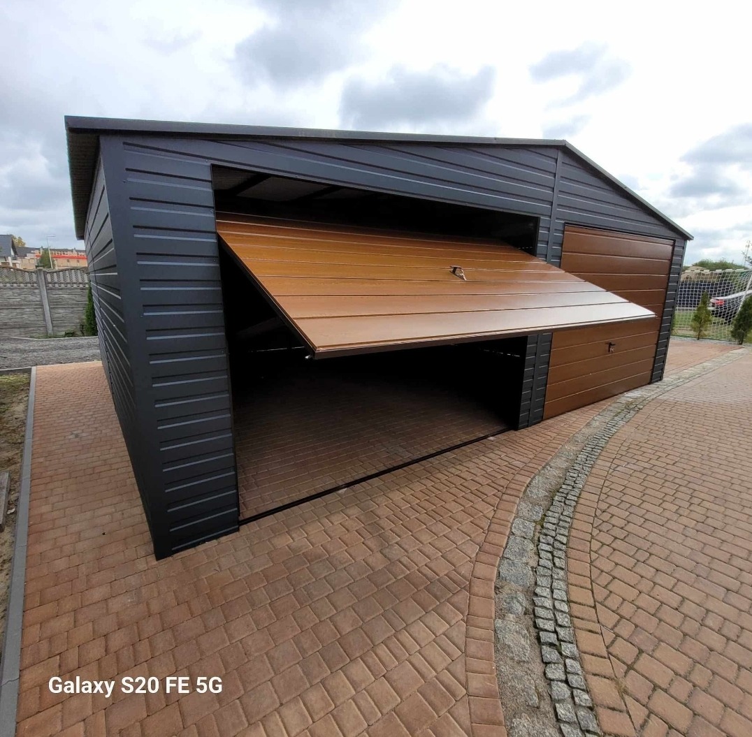 Garaż blaszany z brązową, uchylną bramą w kolorze drewna, częściowo otwartą. Widoczna kostka brukowa i ogrodzenie w tle. Garaż w kolorze grafitowym.