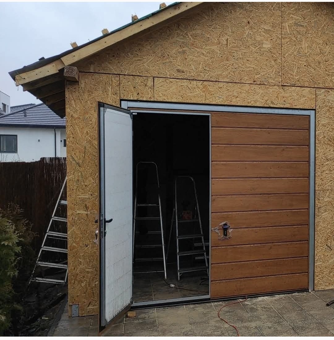 Montaż segmentowej bramy garażowej z drewnianym panelem w garażu z płyty OSB. Wnętrze garażu z drabinami. Widoczny fragment dachu i elewacji domu w tle.