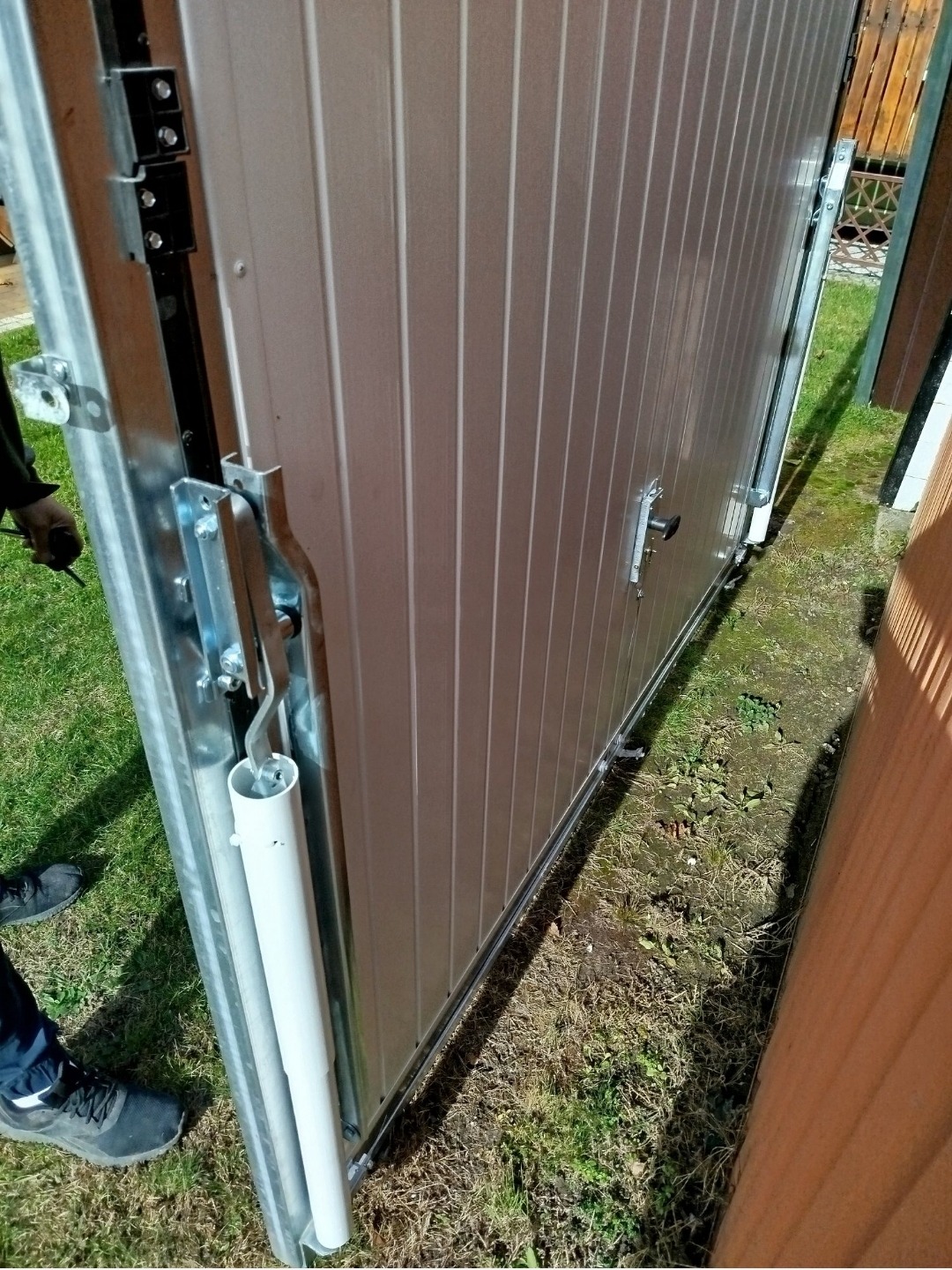 Szczegółowe ujęcie mechanizmu prowadzącego przesuwnej bramy garażowej z widocznymi elementami montażowymi i fragmentem poszycia w kolorze jasnego beżu.