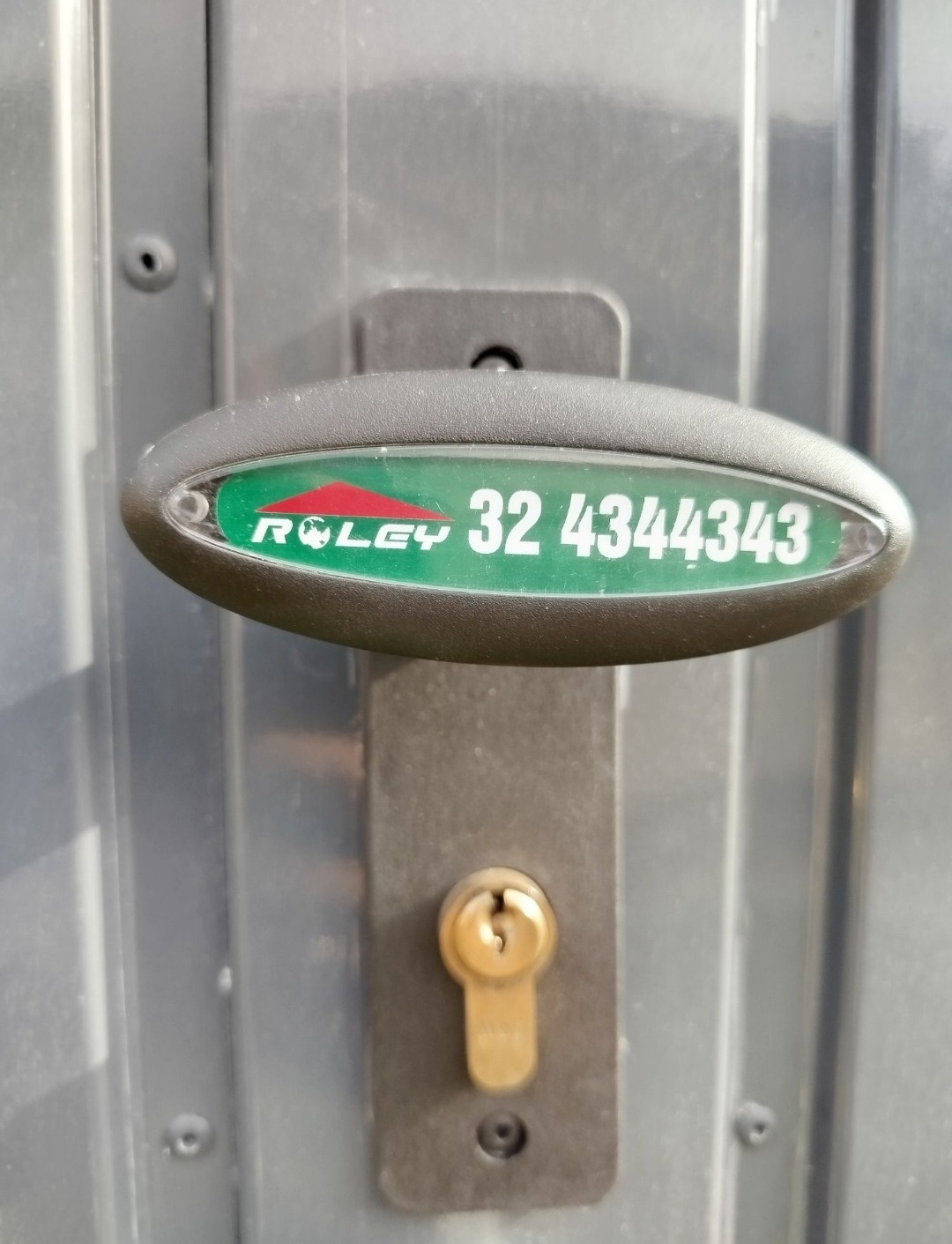Szczegółowe ujęcie zamka i klamki szarej bramy garażowej z naklejką 'ROLEY' i numerem telefonu. Widoczne śruby mocujące i faktura blachy.