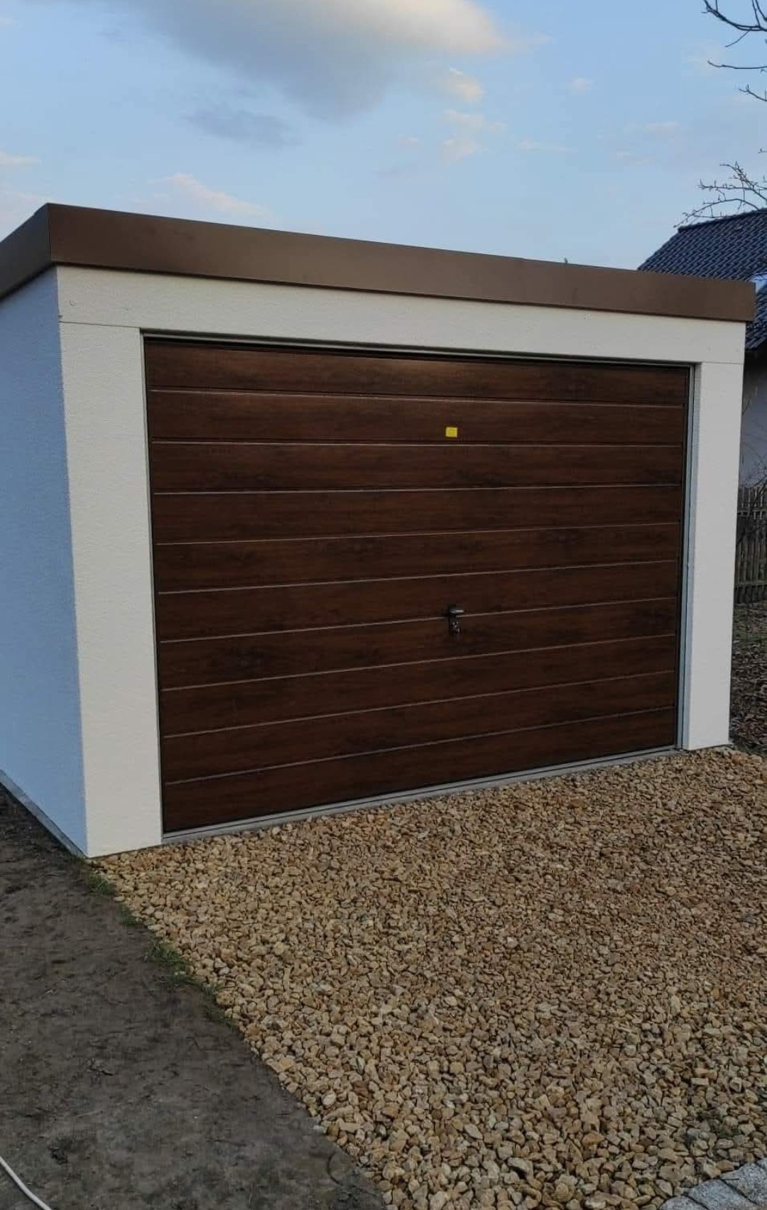 Garaż blaszany z brązową bramą segmentową, wykończony białym obramowaniem i brązowym dachem, otoczony żwirem. Widoczne elementy konstrukcyjne i otoczenie.