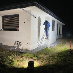 Dawid Grzyb - Praca po zmroku: Elektryk na drabinie montuje oświetlenie zewnętrzne na elewacji domu. Widoczne lampy robocze, narzędzia i fragment elewacji z oknem.