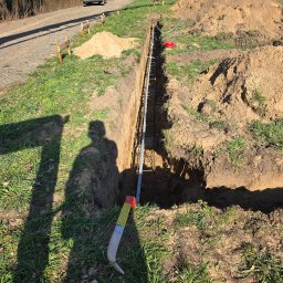 Dawid Grzyb - Wykop zbrojony stalą, przygotowany pod fundament. Widoczny cień osoby i narzędzie. Tło: trawa, ziemia, drzewa, budynki w oddali, błękitne niebo.