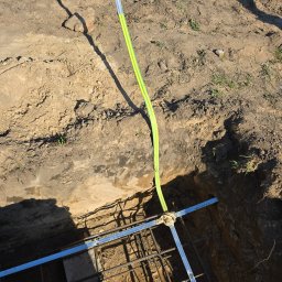 Dawid Grzyb - Wykop z ułożonym zbrojeniem i pionową rurą ochronną z żółto-zielonym paskiem, prawdopodobnie pod instalację odgromową, w słoneczny dzień.