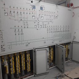 Dawid Grzyb - Rozbudowana tablica elektryczna z otwartymi szafkami, widoczne liczne przewody w żółtej izolacji. Schemat instalacji na ścianie. Elementy automatyki przemysłowej.