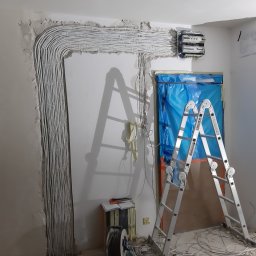 Dawid Grzyb - Białe kable elektryczne ułożone wzdłuż ściany, prowadzące do skrzynki rozdzielczej. Drabina oparta o zabezpieczone folią drzwi. Widoczne narzędzia i przewody na podłodze.