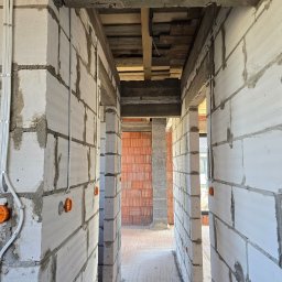 Dawid Grzyb - Wnętrze w trakcie budowy z widocznymi przewodami elektrycznymi i puszkami instalacyjnymi na ścianach z białych bloczków. Widoczne drewniane elementy konstrukcyjne stropu.