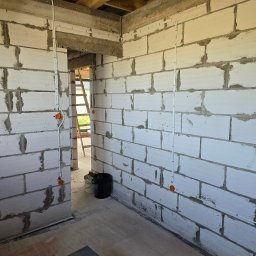 Dawid Grzyb - Wnętrze w trakcie budowy z widoczną instalacją elektryczną w białych rurkach, prowadzoną po ścianach z białych bloczków. Widoczne puszki instalacyjne i drabina w tle.