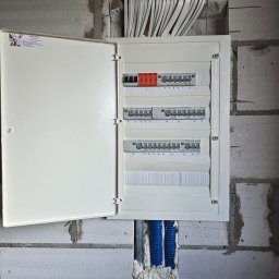 Dawid Grzyb - Otwarta skrzynka elektryczna z bezpiecznikami na ścianie z betonu komórkowego. Widoczne kable i rury instalacyjne. Etykieta z danymi elektryka na drzwiczkach.
