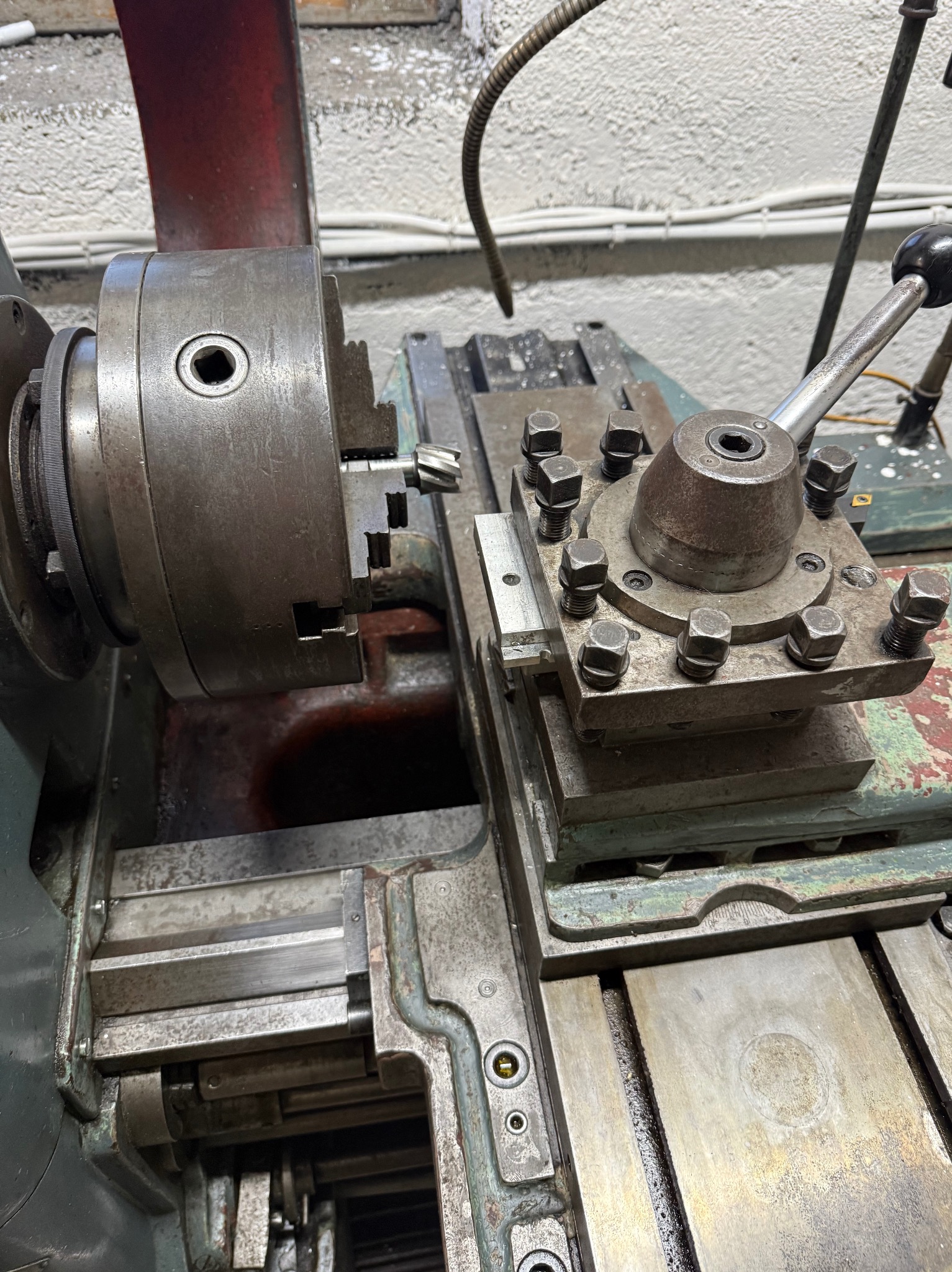 Metalowa tokarka z uchwytem i narzędziem skrawającym, widoczna w warsztacie. Detal maszyny z elementami mocującymi i śladami użytkowania.