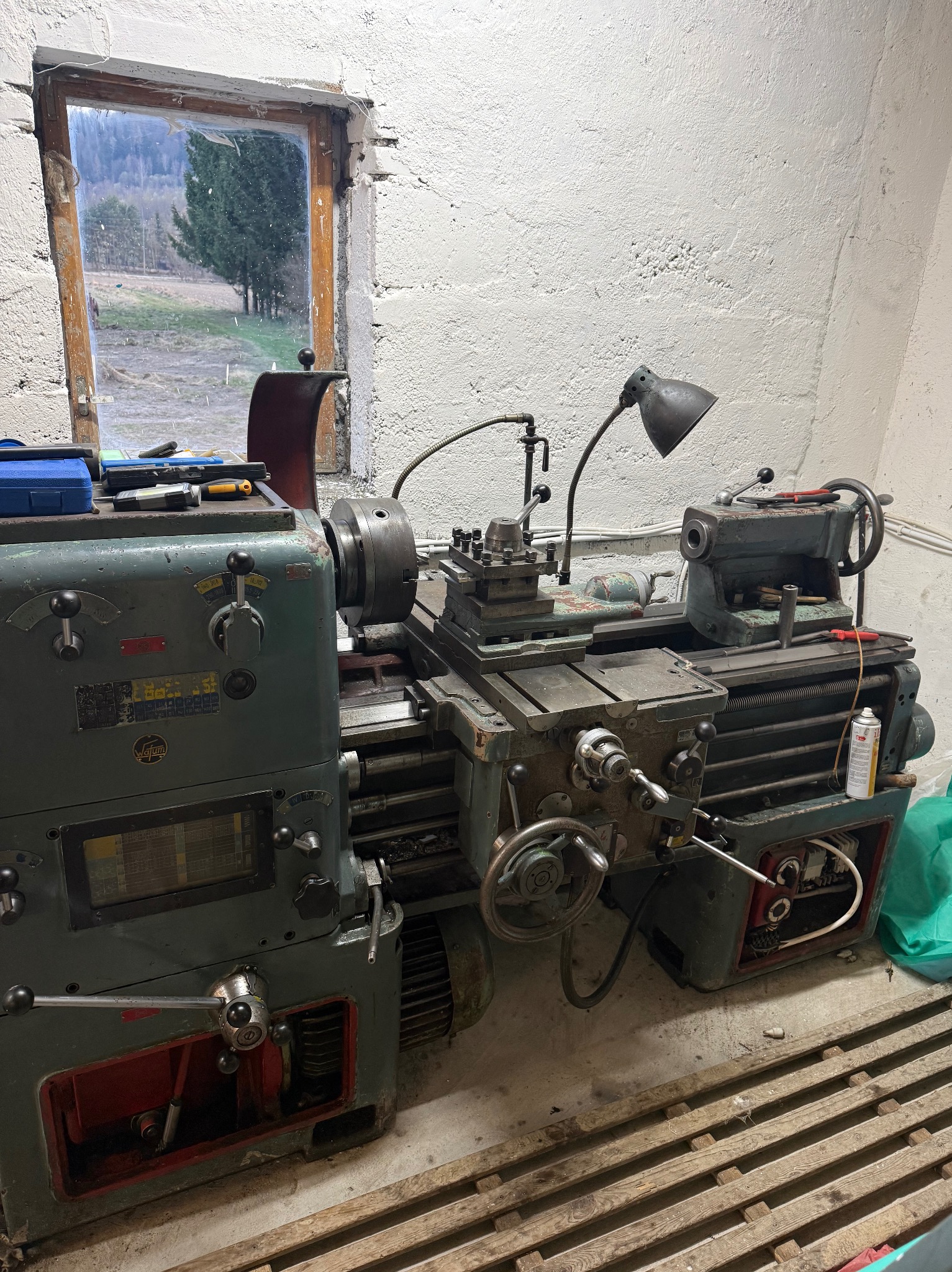 Stare tokarskie urządzenie marki FATUM w warsztacie, oświetlone lampką, z widokiem na zewnątrz przez okno. Widoczne narzędzia i detale maszyny.