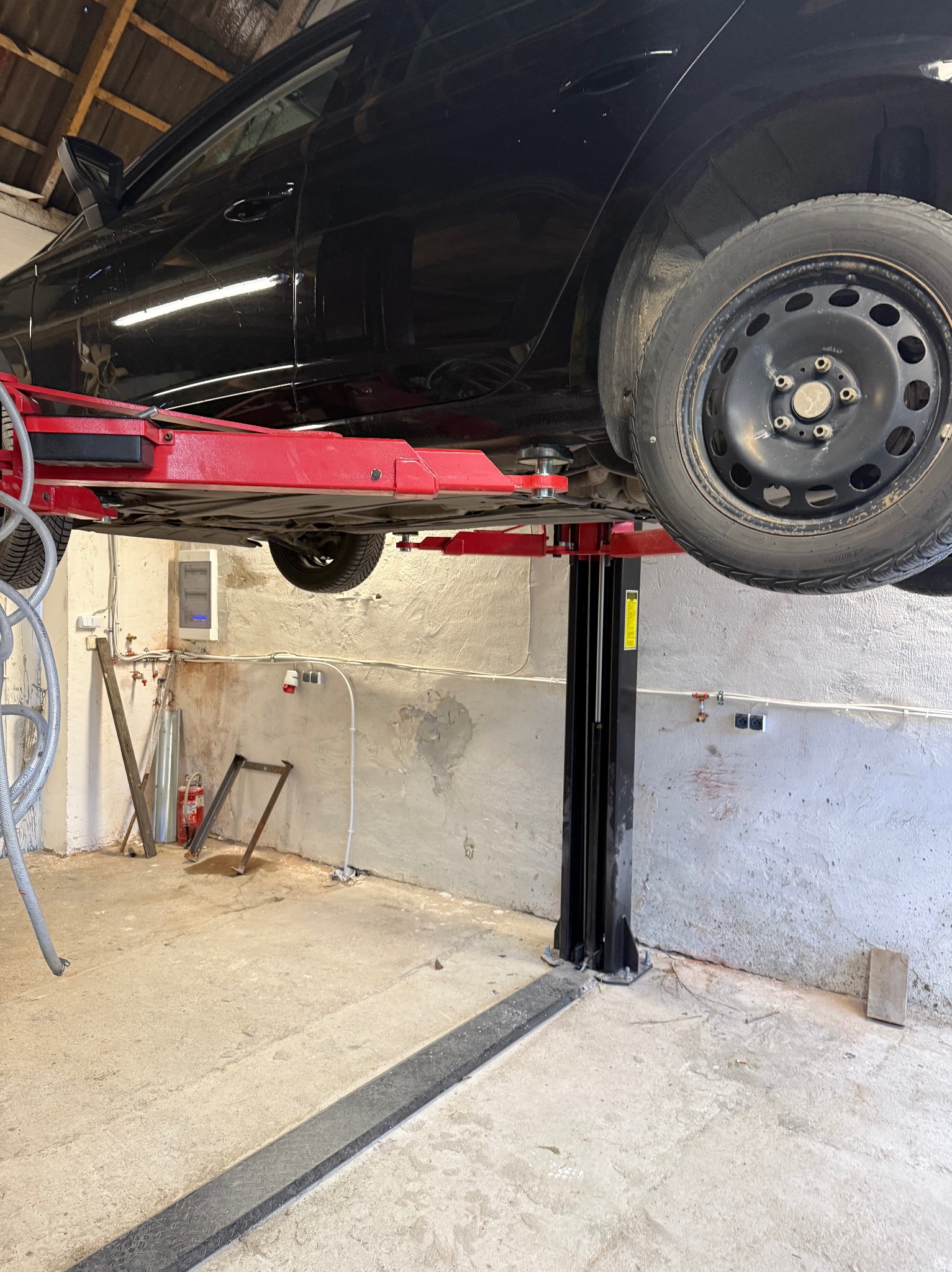 Czarny samochód na podnośniku w warsztacie. Widoczne koło z oponą zimową, podwozie i elementy konstrukcyjne podnośnika. Ściany warsztatu z instalacjami.