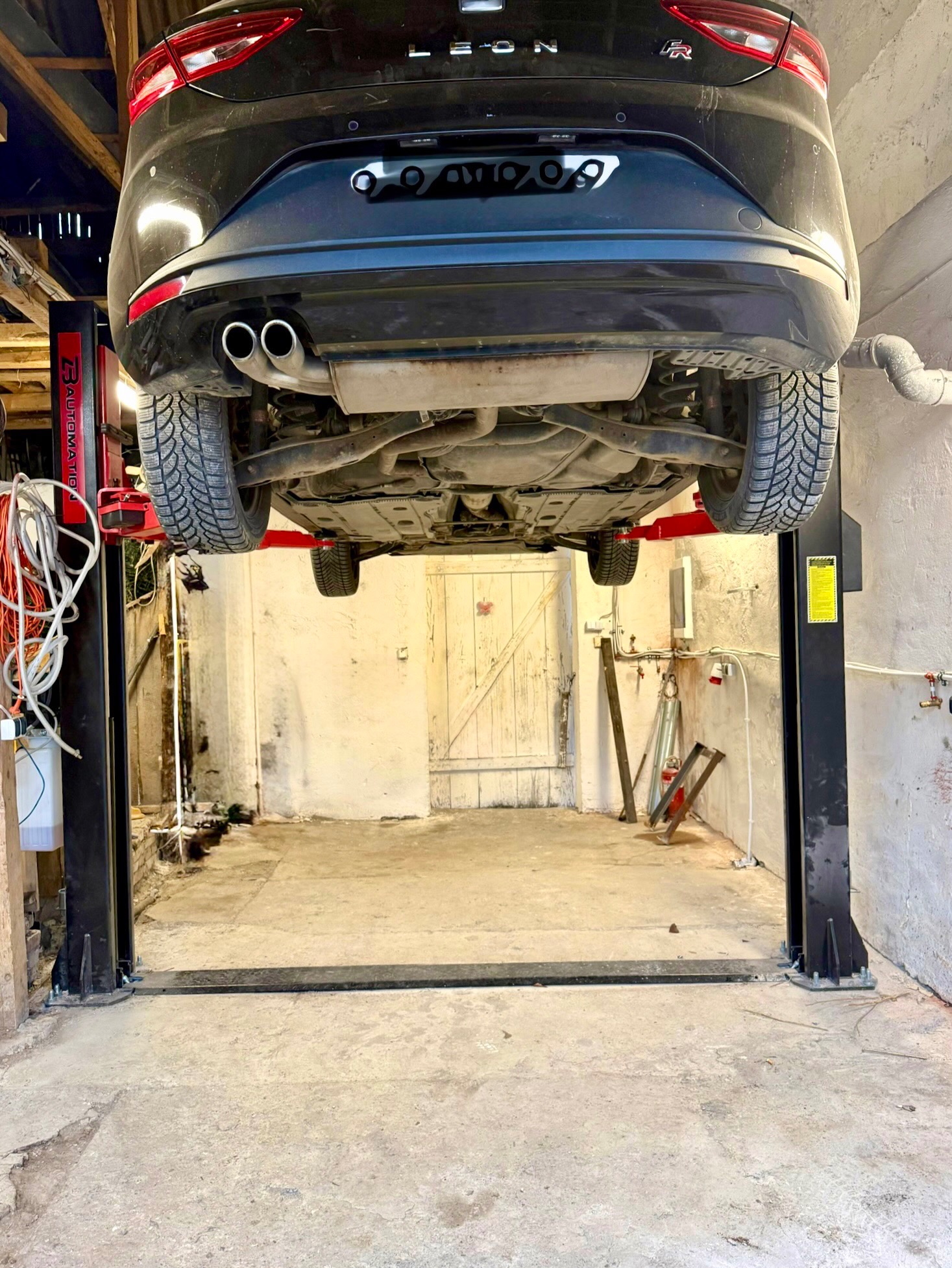 Czarny samochód marki Leon na podnośniku w warsztacie. Widok od spodu, ukazujący zawieszenie, układ wydechowy i opony zimowe. W tle białe ściany i drewniane drzwi.