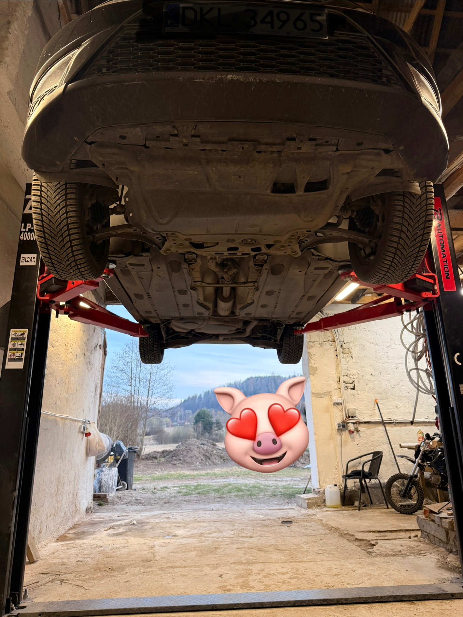 Podwozie auta na podnośniku w warsztacie, widok z dołu. W tle widok na zewnątrz z emoji świnki z sercami zamiast oczu.