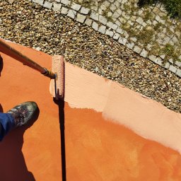 TYZON VICTOR TYSZLEWICZ - Malowanie pomarańczowej powierzchni wałkiem na kiju. Częściowo pomalowana ściana z widoczną fakturą, obok kamienie i kostka brukowa. Buty malarza w lewym dolnym rogu.