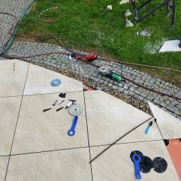 Tyzon Victor Tyszlewicz - Układanie beżowych płytek na zewnątrz w Zabór Mały. Widoczne narzędzia glazurnicze, poziomice i fragment trawnika. Prace w toku, częściowo ukończona powierzchnia.