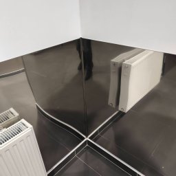 TYZON VICTOR TYSZLEWICZ - Wykończona łazienka z czarnymi, błyszczącymi płytkami, białymi fugami i grzejnikiem. W lustrzanych panelach odbija się sylwetka osoby. Nowoczesny design.