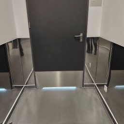 Tyzon Victor Tyszlewicz - Nowoczesne wykończenie korytarza z ciemną podłogą, metalowymi listwami przypodłogowymi i drzwiami. Widoczne odbicie osoby w metalowych elementach.