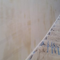 TYZON VICTOR TYSZLEWICZ - Przygotowana ściana do malowania z widocznymi plamami i zabezpieczone schody płachtą malarską z niebieską taśmą. Widok z dołu.