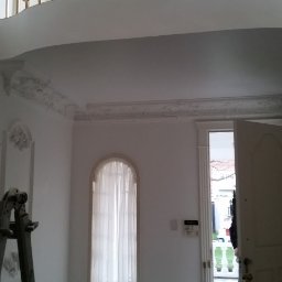 Tyzon Victor Tyszlewicz - Białe wnętrze z balkonem i dekoracyjnymi sztukateriami. Widoczna drabina malarska, okno z firanką i otwarte drzwi. Marmurowa podłoga i złote zdobienia balustrady.