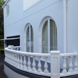 TYZON VICTOR TYSZLEWICZ - Biała fasada budynku z balkonem o balustradzie z tralkami. Widoczne okna łukowe i drzwi. W tle zieleń drzew i krzewów. Świeżo pomalowana elewacja.