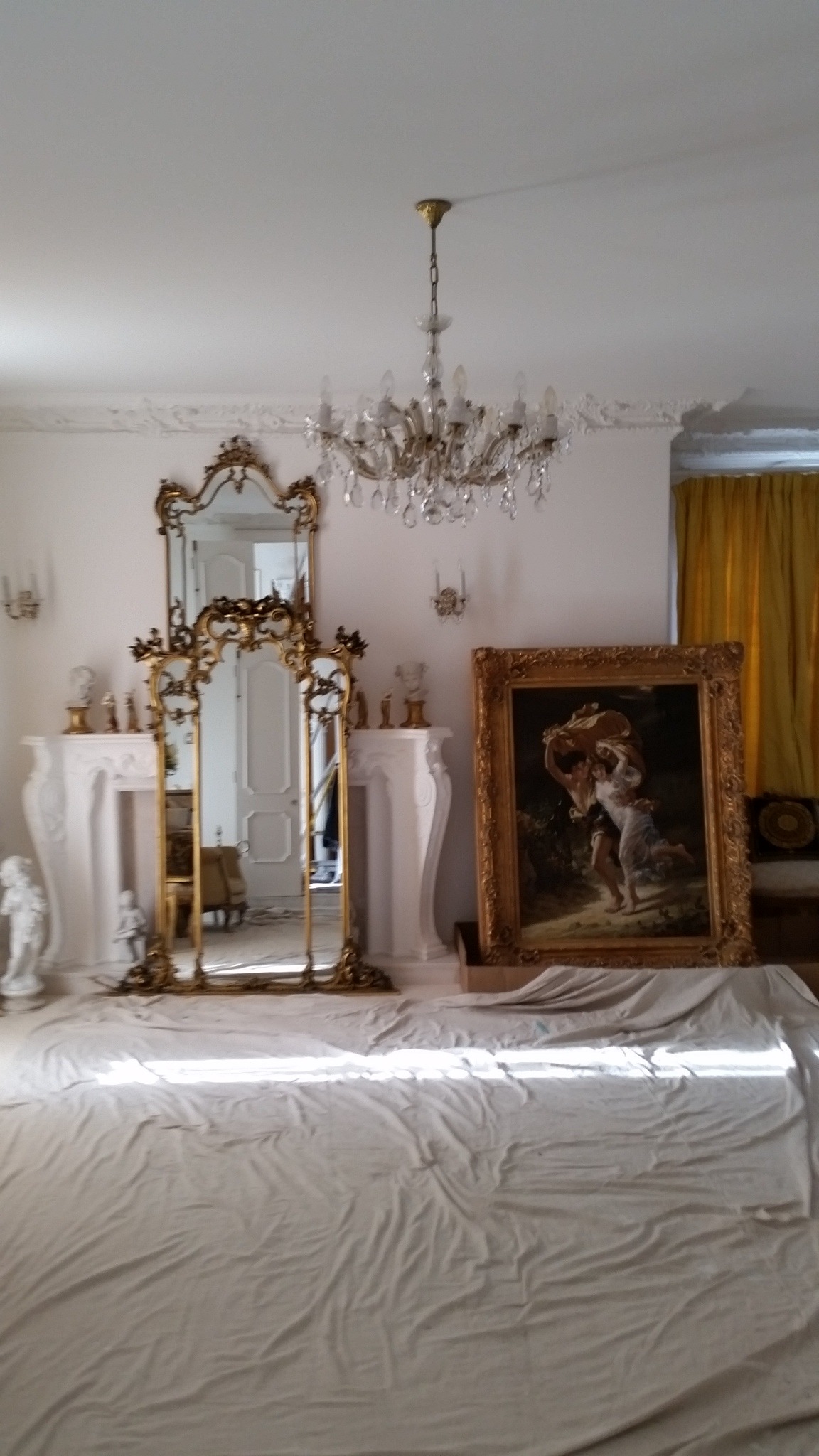 Pokój przed remontem: zabezpieczone meble pod płachtą, dwa lustra w złoconych ramach, obraz w złotej ramie, żyrandol kryształowy, dekoracyjny gzyms.