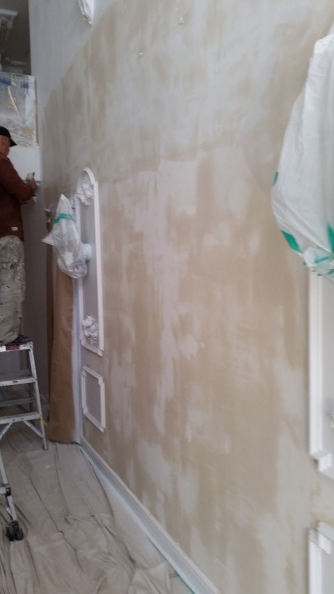 Prace malarskie w trakcie realizacji w Zabór Mały. Widoczna ściana z dekoracyjnymi elementami, malarz na drabinie i zabezpieczona podłoga.