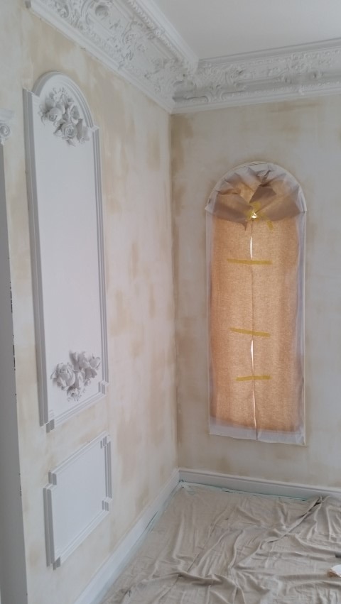 Pokój w trakcie prac wykończeniowych: ściany z dekoracyjnymi sztukateriami, okno zabezpieczone folią, podłoga przykryta płachtą malarską. Przygotowanie do malowania.