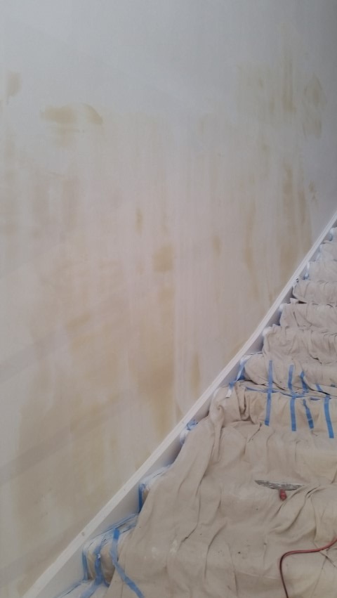 Przygotowana ściana do malowania z widocznymi plamami i zabezpieczone schody płachtą malarską z niebieską taśmą. Widok z dołu.