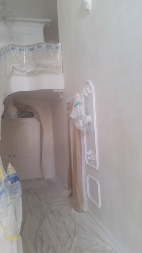 Wnętrze po malowaniu na biało, z dekoracyjnymi sztukateriami na ścianie i zabezpieczoną folią podłogą. Widoczny fragment balkonu i drzwi.