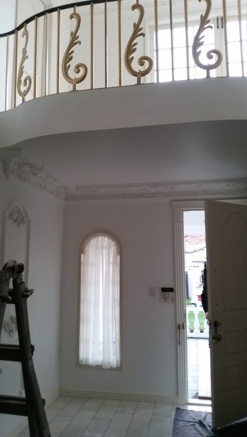 Białe wnętrze z balkonem i dekoracyjnymi sztukateriami. Widoczna drabina malarska, okno z firanką i otwarte drzwi. Marmurowa podłoga i złote zdobienia balustrady.