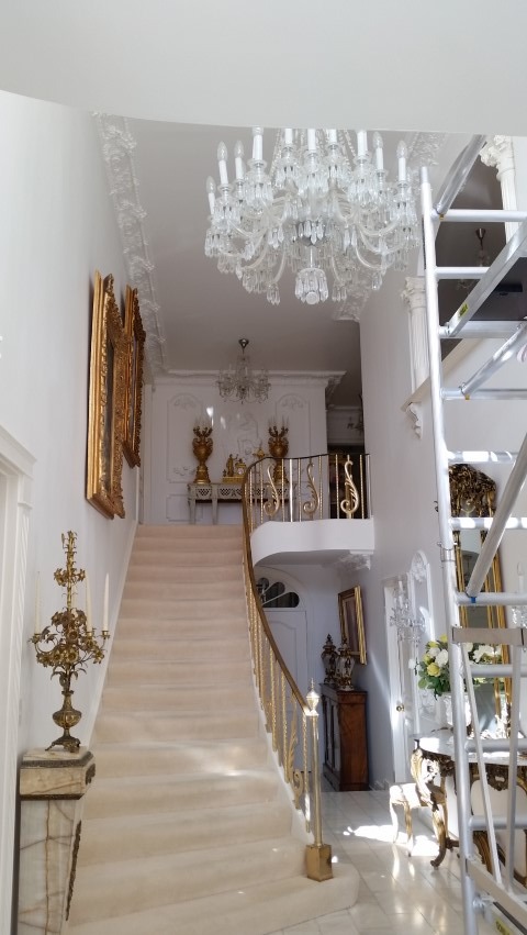 Jasne wnętrze z marmurowymi schodami i kryształowymi żyrandolami. Widoczne rusztowanie malarskie, sugerujące prace wykończeniowe w eleganckim domu.