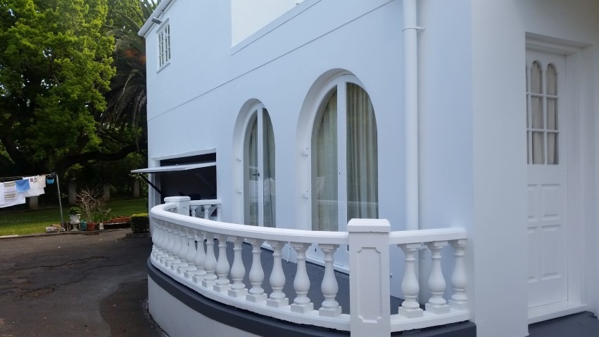 Biała fasada budynku z balkonem o balustradzie z tralkami. Widoczne okna łukowe i drzwi. W tle zieleń drzew i krzewów. Świeżo pomalowana elewacja.