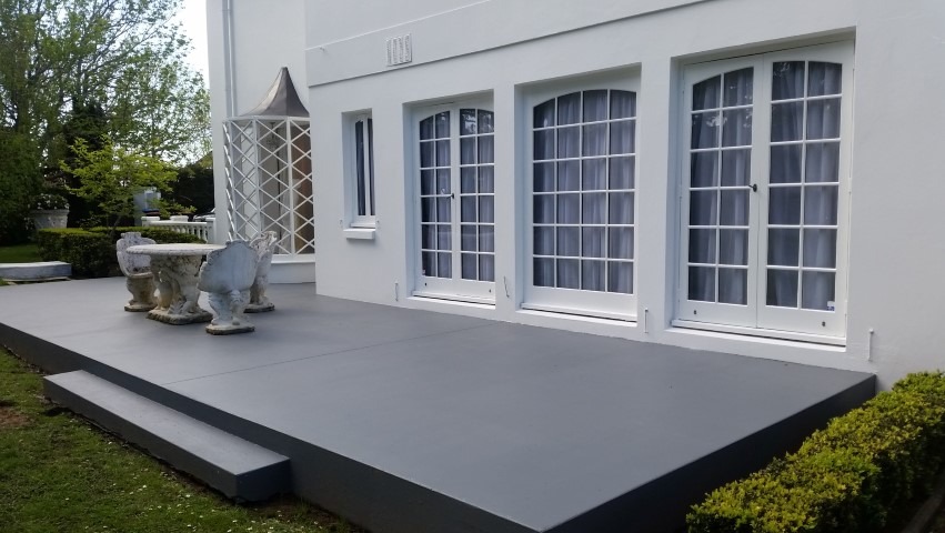 Szary taras z białymi drzwiami balkonowymi i kamiennym stołem z krzesłami. Widok na zadbany ogród. Minimalistyczny design.