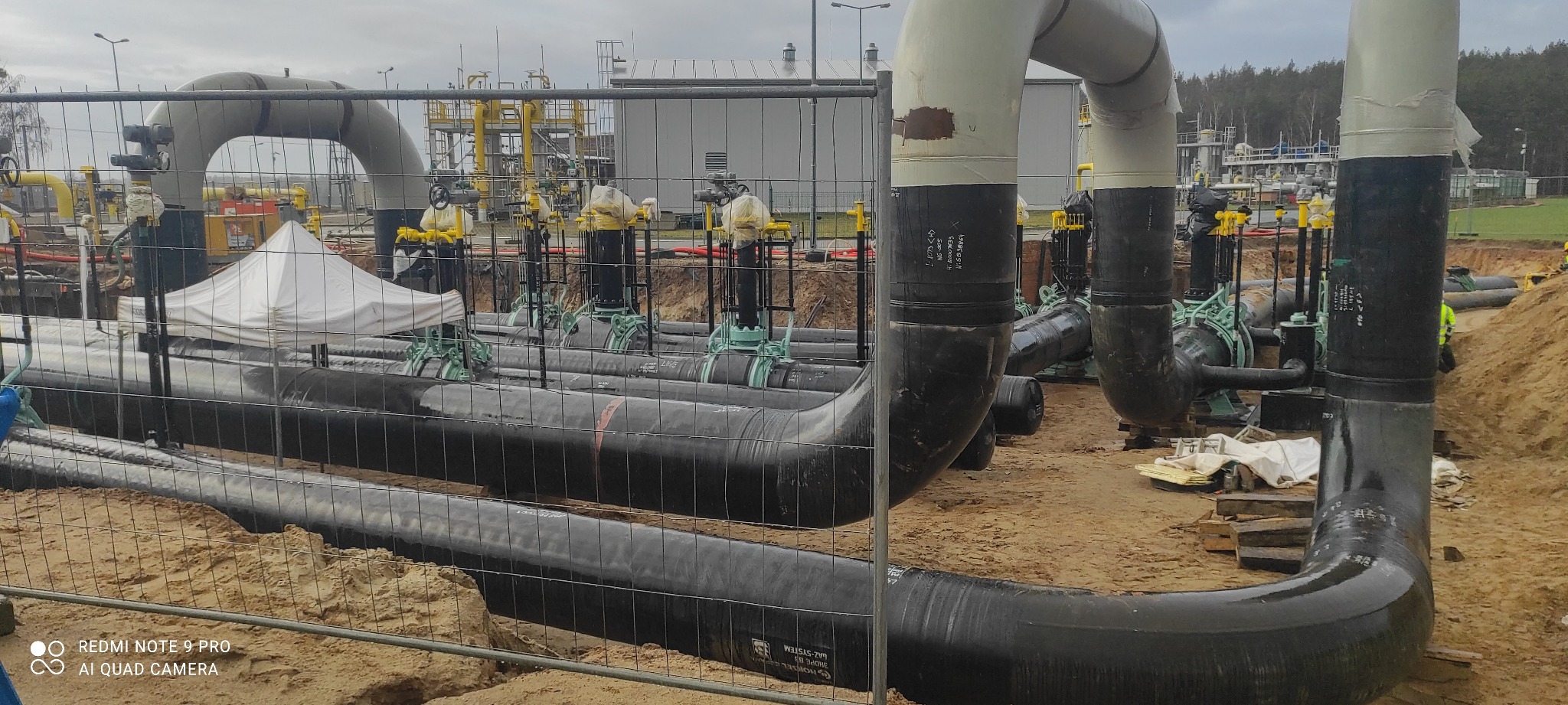 Rury gazowe w wykopie, zabezpieczone siatką. Widoczne elementy instalacji, zawory i fragmenty konstrukcji. Prace ziemne w toku, teren budowy.