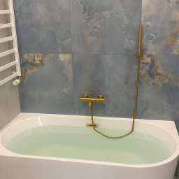 Primebud - Wanna wypełniona wodą, z baterią w kolorze starego złota i grzejnikiem drabinkowym. Tło stanowi ściana wyłożona płytkami imitującymi marmur w odcieniach błękitu i brązu.