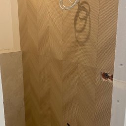 Primebud - Ukończona ściana w łazience z beżowymi płytkami ułożonymi w jodełkę. Widoczne przygotowane punkty instalacji i wystające przewody. Fragment podłogi z płytek.