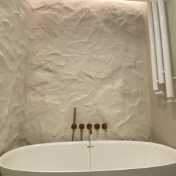 Primebud - Nowoczesna łazienka z wolnostojącą wanną, armaturą w kolorze złota i ścianą o fakturze kamienia, podświetloną od góry. Minimalistyczny grzejnik drabinkowy.