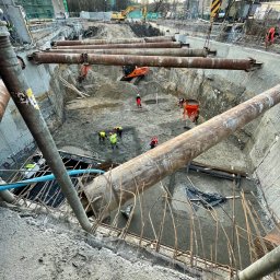 FRONT Budownictwo Kamil Zborowski - Głęboki wykop zabezpieczony stalowymi rurami, wewnątrz pracownicy i sprzęt budowlany. Widok z góry na plac budowy w trakcie realizacji.