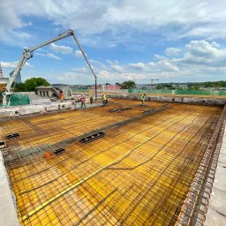 FRONT Budownictwo Kamil Zborowski - Zbrojenie stropu przygotowane do zalewania betonem. Widoczna pompa do betonu i pracownicy w kaskach. Żółte szalunki i stalowe pręty tworzą siatkę. W tle zabudowa miejska.