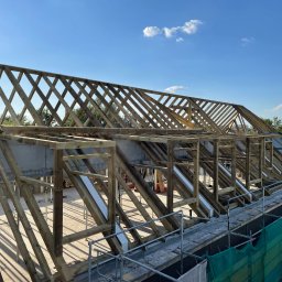 FRONT Budownictwo Kamil Zborowski - Konstrukcja drewniana dachu w trakcie budowy, widok z góry na rusztowanie, błękitne niebo w tle. Elementy zaimpregnowane, widoczne detale łączeń.