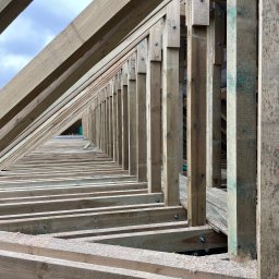 FRONT Budownictwo Kamil Zborowski - Konstrukcja drewniana dachu w trakcie budowy, widok z dołu. Elementy połączone śrubami, widoczne wióry. Perspektywa zbieżna w głąb konstrukcji.