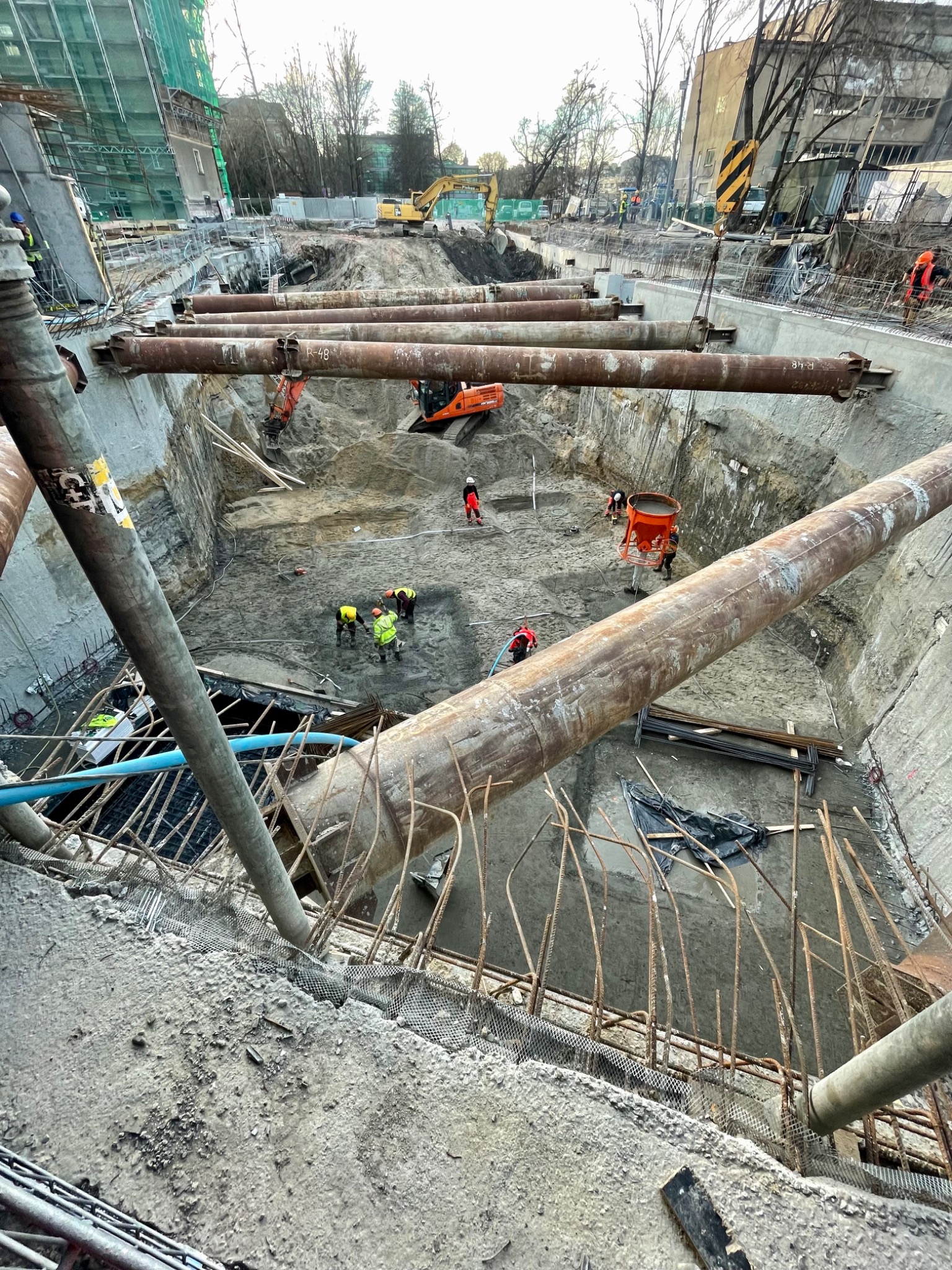 Głęboki wykop zabezpieczony stalowymi rurami, wewnątrz pracownicy i sprzęt budowlany. Widok z góry na plac budowy w trakcie realizacji.