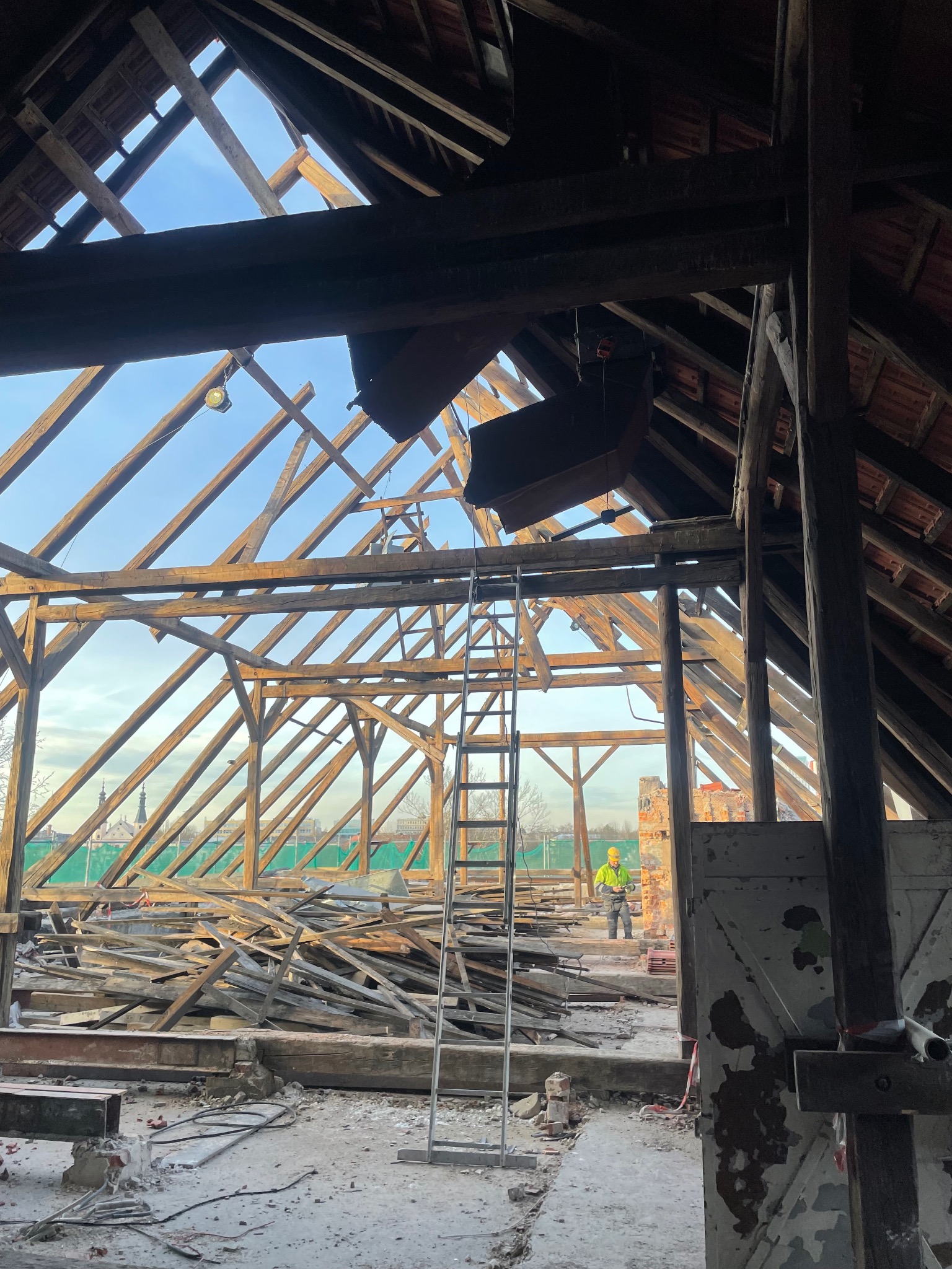 Demontaż drewnianej konstrukcji dachu z widoczną drabiną i pracownikiem w kasku, w tle zabudowa miejska. Prace rozbiórkowe wewnątrz budynku.