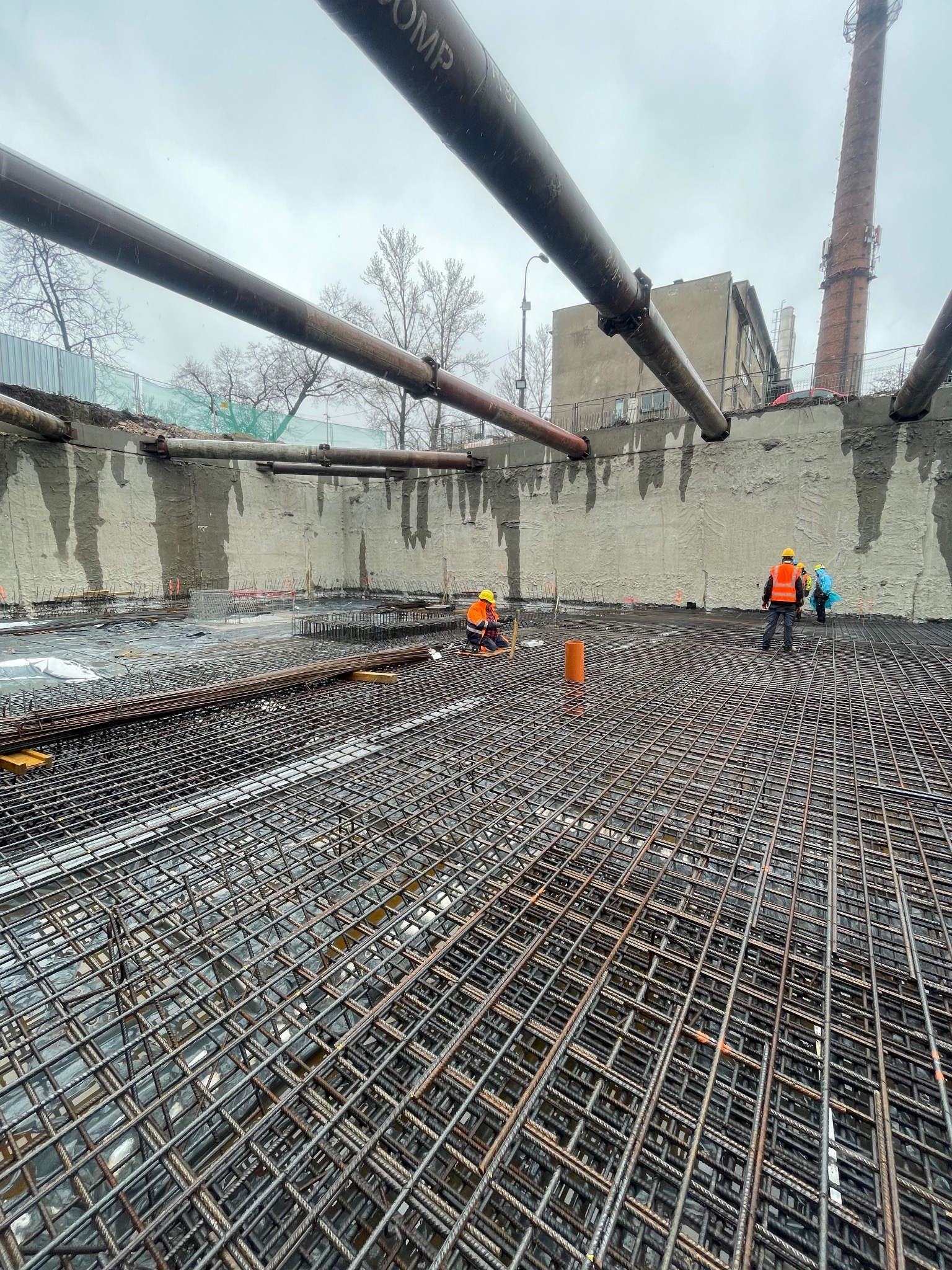 Zbrojenie płyty fundamentowej z widocznymi pracownikami w odblaskowych kamizelkach. W tle betonowa ściana i komin. Widok na siatkę zbrojeniową z bliska.