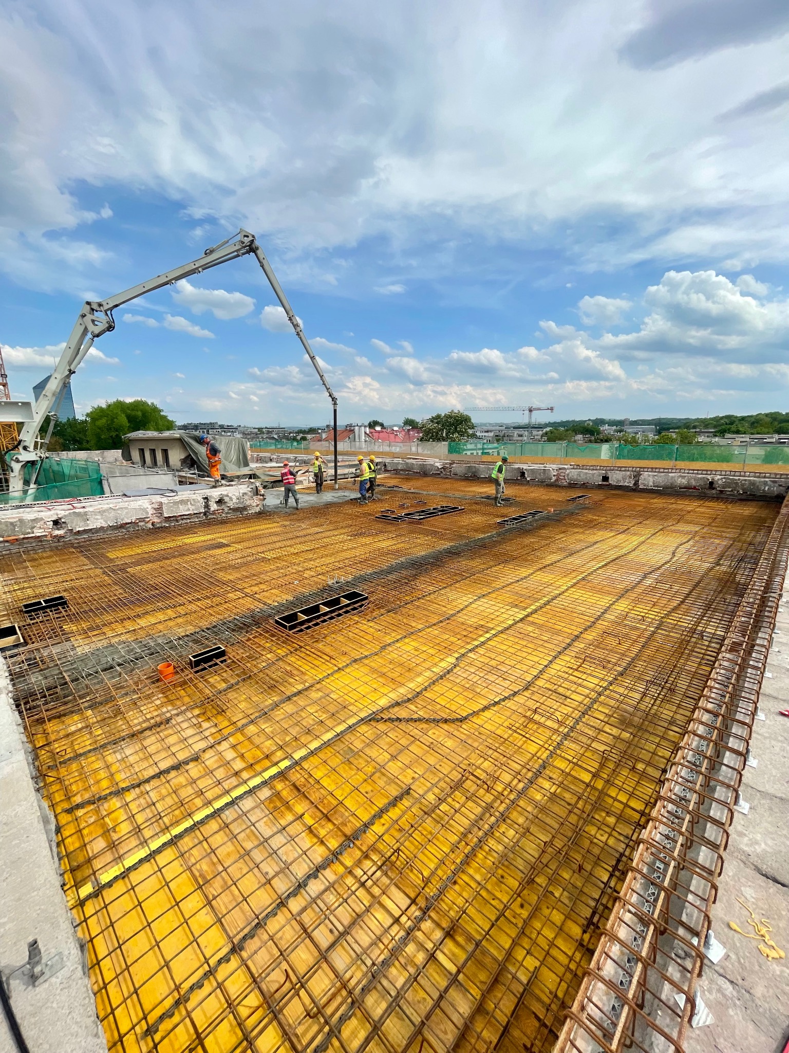 Zbrojenie stropu przygotowane do zalewania betonem. Widoczna pompa do betonu i pracownicy w kaskach. Żółte szalunki i stalowe pręty tworzą siatkę. W tle zabudowa miejska.