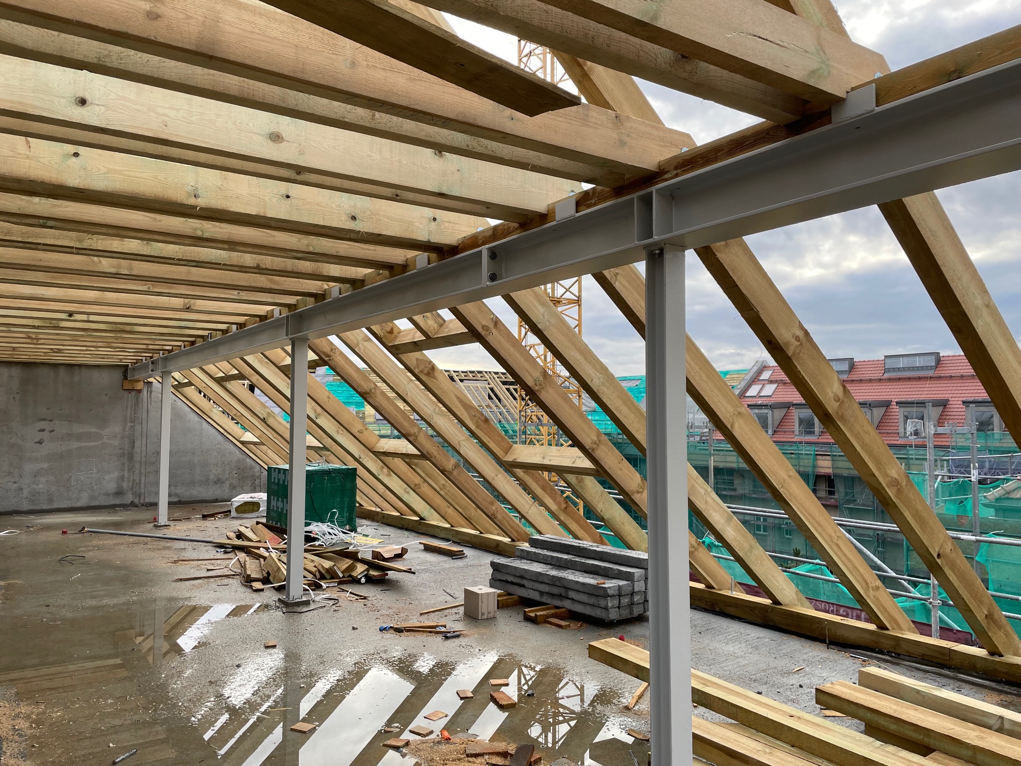 Konstrukcja dachu z drewnianych belek i stalowych podpór w surowym stanie, widok na budynek w tle i odbicia w wodzie na podłodze.