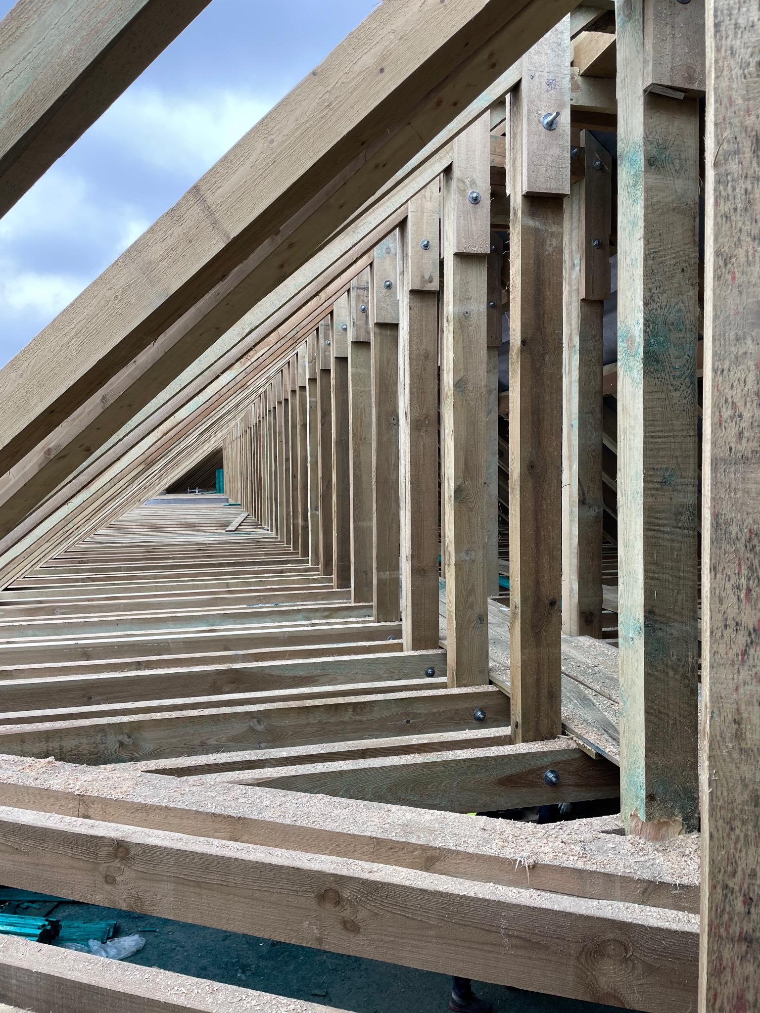 Konstrukcja drewniana dachu w trakcie budowy, widok z dołu. Elementy połączone śrubami, widoczne wióry. Perspektywa zbieżna w głąb konstrukcji.