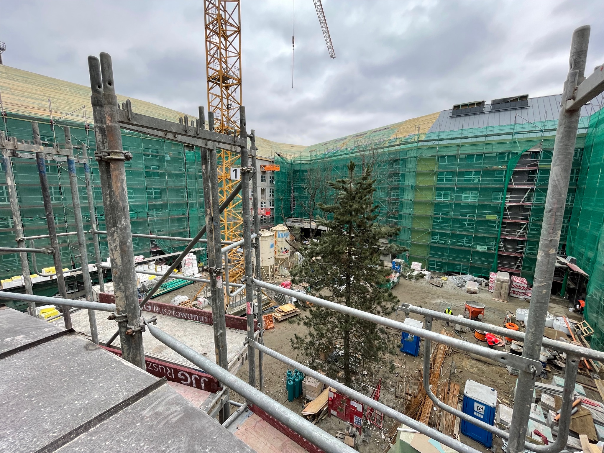Budynek w trakcie budowy, widok z rusztowania. Siatka ochronna, żuraw budowlany i drzewo na placu budowy. Widok z góry na teren budowy.