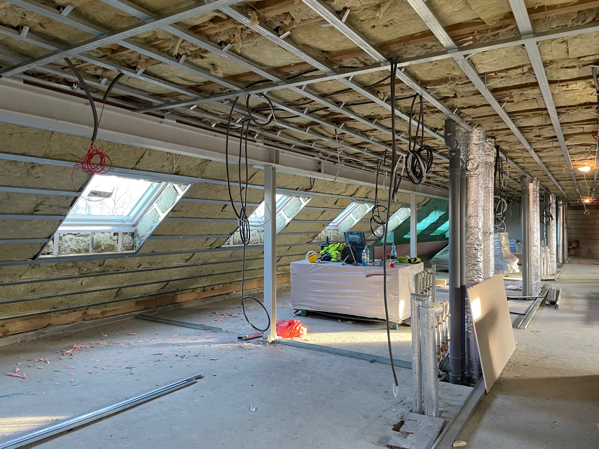 Ocieplenie poddasza wełną mineralną, widoczne okna dachowe, metalowa konstrukcja sufitu podwieszanego i instalacje. Prace wykończeniowe w surowym wnętrzu.